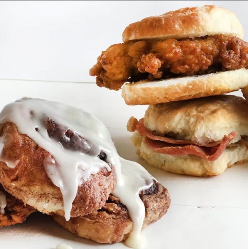 A cinnamon roll with icing beside a biscuit sandwich filled with fried chicken and another with ham.