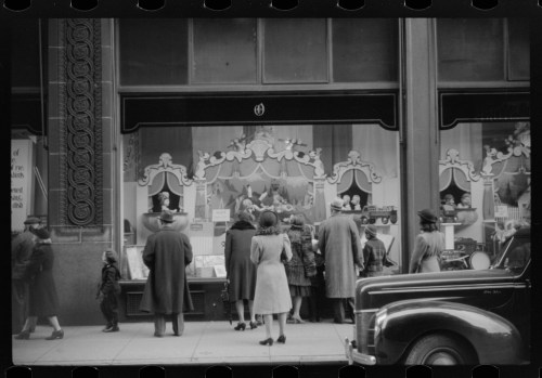 A group of people, dressed in vintage clothing, admire a store window display featuring toys and decorations.