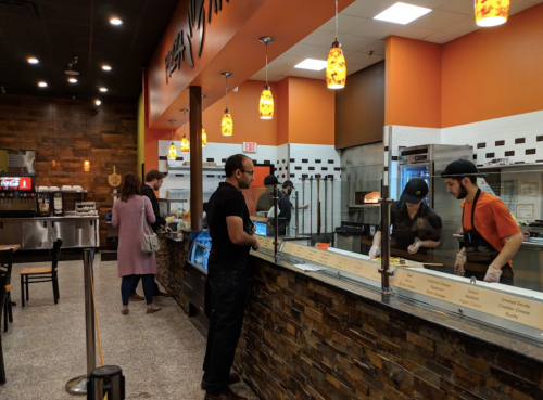 A modern restaurant interior with customers ordering at the counter and staff preparing food behind a glass barrier.
