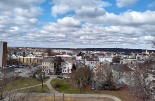 A panoramic view of a town with various buildings, trees, and a cloudy sky in the background.