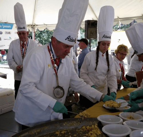 A group of chefs in white hats prepares food at an outdoor event, serving dishes to attendees under a tent.
