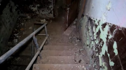 A dimly lit staircase leading down, surrounded by peeling paint and debris, with a metal railing on the side.