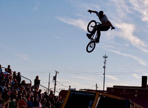 A BMX rider performs a mid-air trick above a crowd during a stunt show against a clear sky.