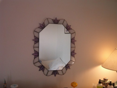 A decorative mirror with a floral design hangs on a wall beside a lamp and a bedside table.
