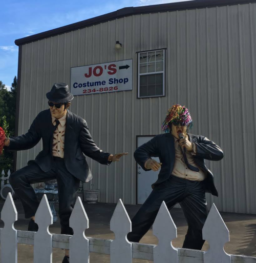 Two life-sized statues of men in suits and hats, one holding a microphone, outside Jo's Costume Shop.