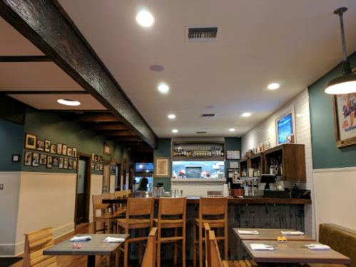 Interior of a cozy restaurant featuring wooden tables, bar stools, and a well-lit counter with shelves and framed photos.