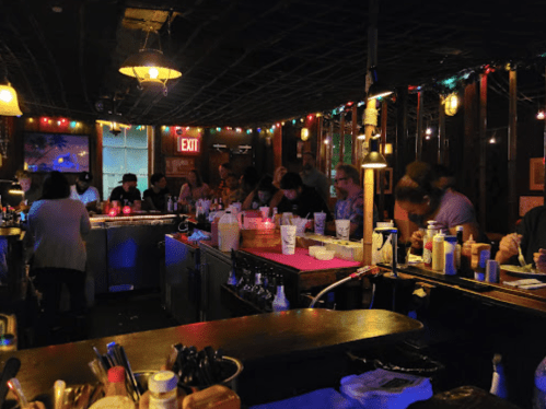 A dimly lit bar scene with patrons seated at the counter, colorful lights, and a lively atmosphere.