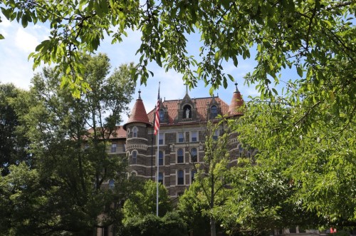 Historic building with turrets, surrounded by green trees and an American flag in front, under a clear blue sky.