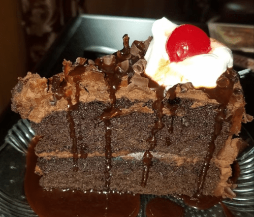 A slice of rich chocolate cake topped with whipped cream and a cherry, drizzled with chocolate sauce.