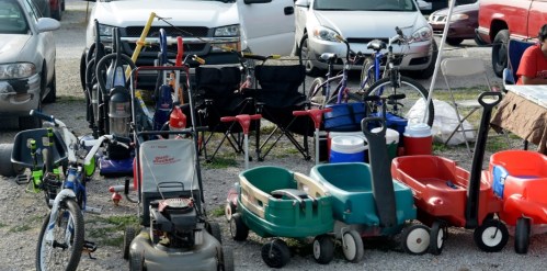 A collection of various items for sale, including bicycles, lawn equipment, coolers, and wagons, set up in a parking area.