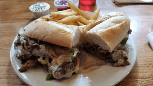 A plate with two cheesesteak sandwiches, topped with onions and peppers, served with a side of fries and dipping sauces.