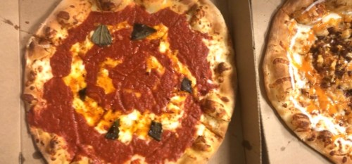 A pizza with a red sauce design and basil leaves, placed in a pizza box next to another pizza.
