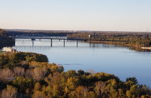 A serene river scene with a bridge, surrounded by trees in autumn colors under a clear sky.