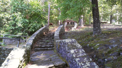 A winding stone path leads through a wooded area, with steps and moss-covered walls surrounded by trees.