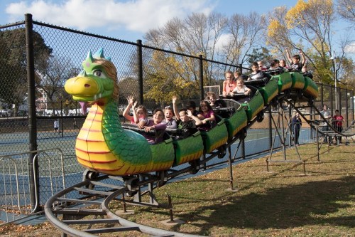 A colorful dragon-themed roller coaster with excited riders waving, set in a park with trees and a fence in the background.