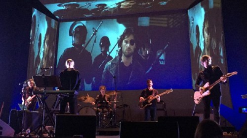 A band performs on stage with a large video projection in the background, featuring musicians and dramatic visuals.