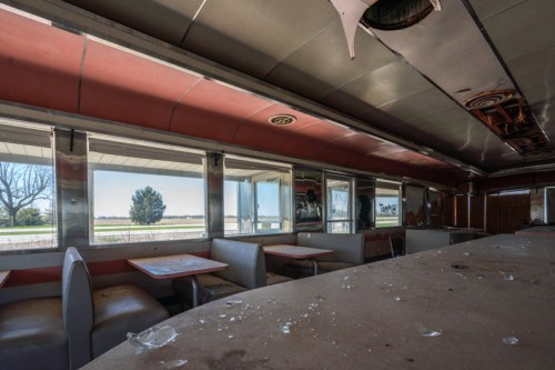 Interior of an abandoned diner with empty booths, shattered glass, and large windows showing a view of the outside.