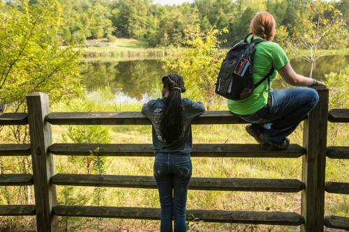 Two people by a wooden fence overlooking a serene pond surrounded by trees. One sits on the fence, the other leans against it.