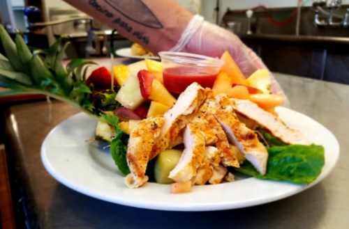 A plate of salad topped with grilled chicken, mixed fruits, and a small cup of dressing, held by a gloved hand.