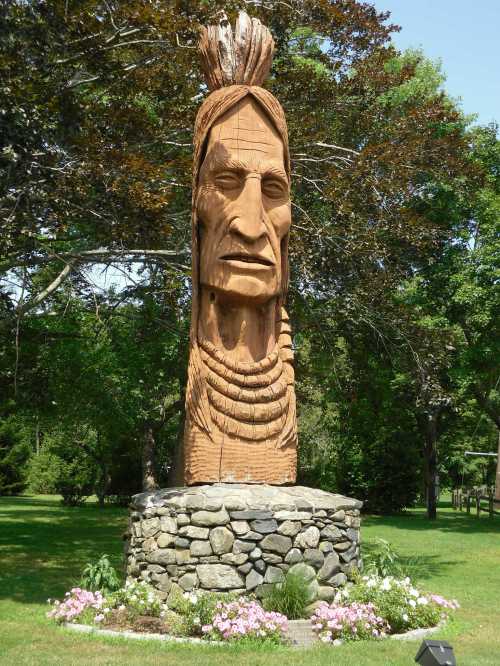 A tall wooden sculpture of a Native American figure, surrounded by flowers and trees in a grassy area.