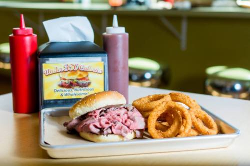 A plate with a roast beef sandwich on a bun and crispy onion rings, with condiments in the background.