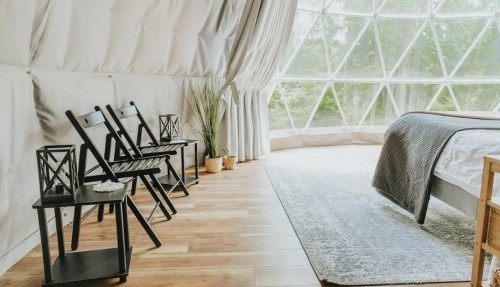 Cozy interior of a geodesic dome with a bed, wooden floor, and minimalist furniture, surrounded by large windows.