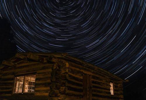 A log cabin under a starry night sky, with star trails creating circular patterns around it.
