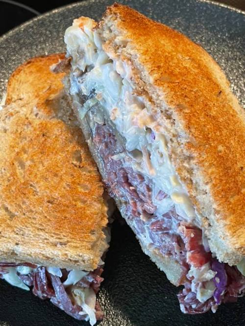 A toasted sandwich filled with layers of meat and creamy coleslaw, cut in half and served on a dark plate.