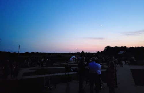 A twilight scene with people gathered outdoors, silhouetted against a colorful sky and distant horizon.