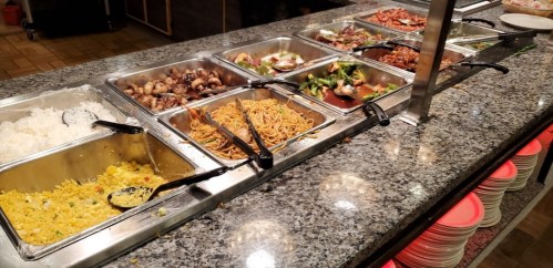 A buffet spread featuring various dishes, including rice, noodles, vegetables, and meats, on a granite countertop.