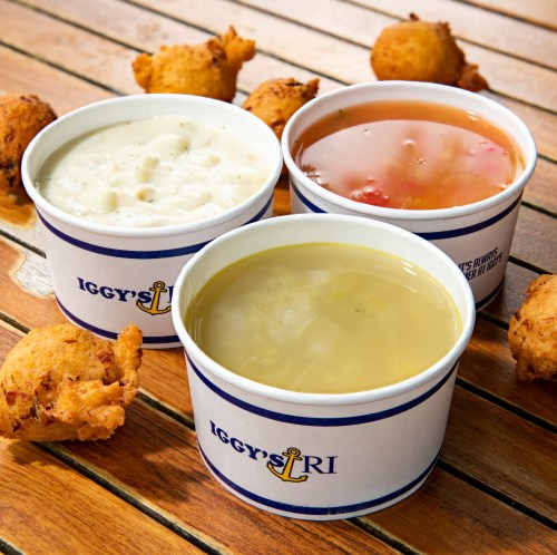 Three cups of soup—clam chowder, tomato soup, and a yellow broth—surrounded by golden fried fritters on a wooden table.