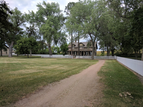 A pathway leads to a large house surrounded by trees and a white picket fence on a grassy lawn.