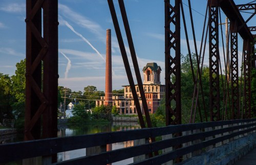A historic factory building and smokestack are seen through a metal bridge, with trees and a river in the foreground.