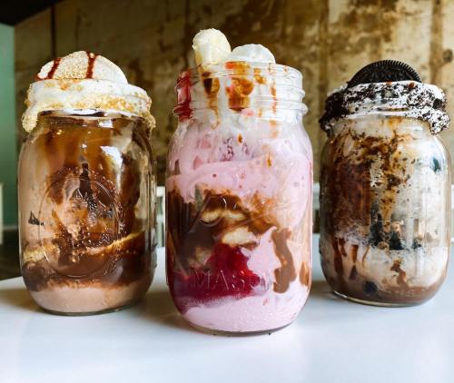Three mason jars filled with colorful ice cream sundaes topped with whipped cream, cookies, and chocolate syrup.
