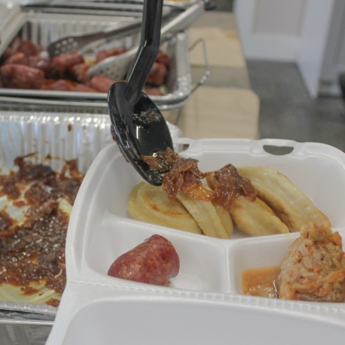A takeout container with sausage, dumplings, and sauce, with a serving spoon in the foreground.