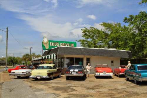 A vintage drive-in restaurant with classic cars parked outside under a blue sky and green trees.