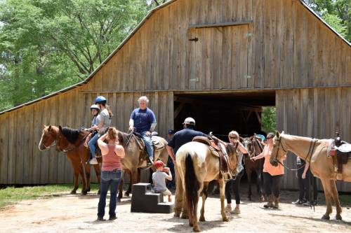 A group of people preparing for a horseback ride near a wooden barn, with children and adults interacting with horses.