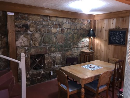 Cozy basement dining area with a wooden table, chairs, stone wall, and a lamp, featuring warm lighting.