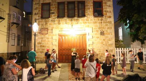 A group of people gathers outside a stone building at night, with a large wooden door and street lamps illuminating the scene.