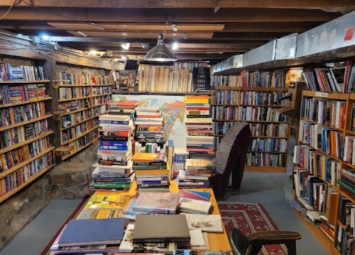 A cozy basement library filled with bookshelves, stacks of books, and a table covered in various books and a map.