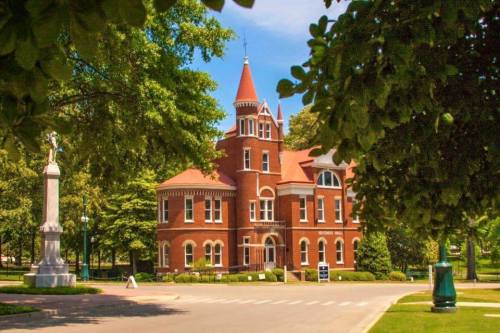Historic red brick building with a turret, surrounded by trees and a clear blue sky. A monument stands nearby.