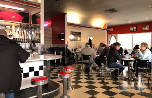 A retro diner interior with checkered floors, patrons seated at tables, and a counter with drink dispensers.