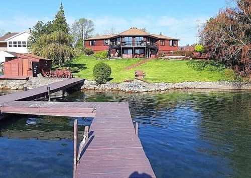 A wooden dock leads to a serene lake with a large house and green lawn in the background under a clear blue sky.