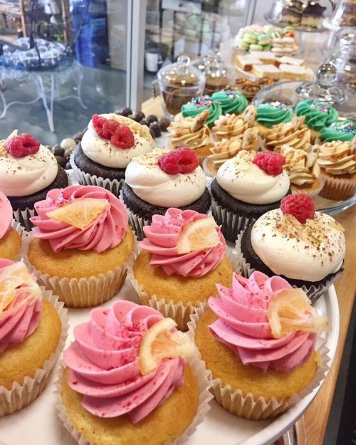 A display of colorful cupcakes with pink and white frosting, topped with fruit, alongside various other desserts.