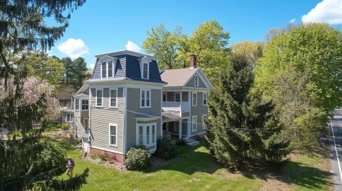 A large, two-story gray house surrounded by trees and greenery on a sunny day.