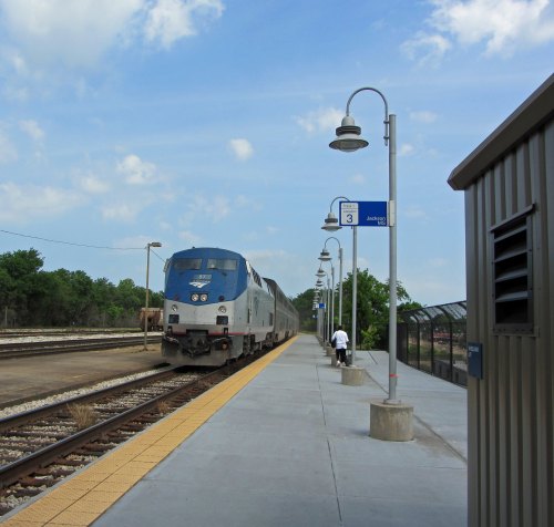 A train at a station with a passenger walking on the platform under blue skies and street lamps.
