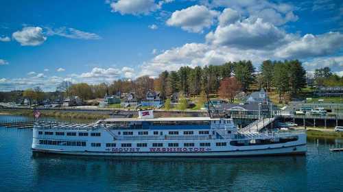 A large white boat named "Mount Washington" docked by a scenic shoreline with trees and houses under a blue sky.