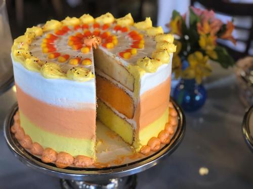 A colorful layered cake with orange, yellow, and white frosting, partially sliced, on a silver cake stand.