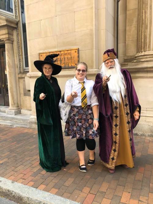 Three people dressed as wizarding characters stand outside a building, holding wands and posing for the photo.
