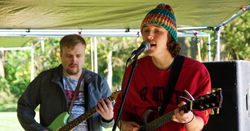 Two musicians perform outdoors; one sings and plays guitar, while the other plays a green electric guitar.